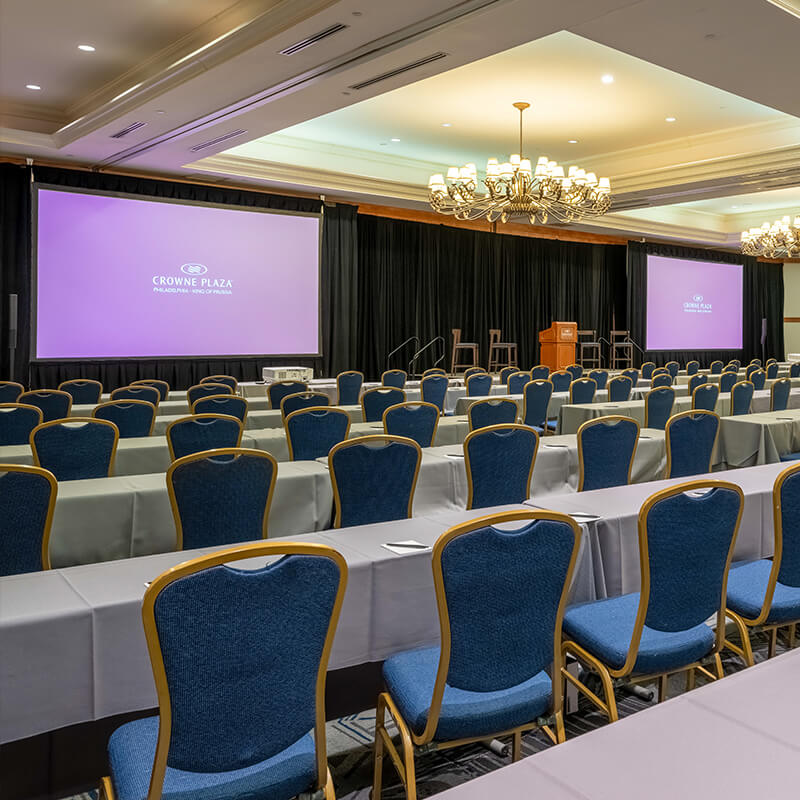 Ballroom with long gray tables and blue chairs and 2 large projector screens at the from of the room