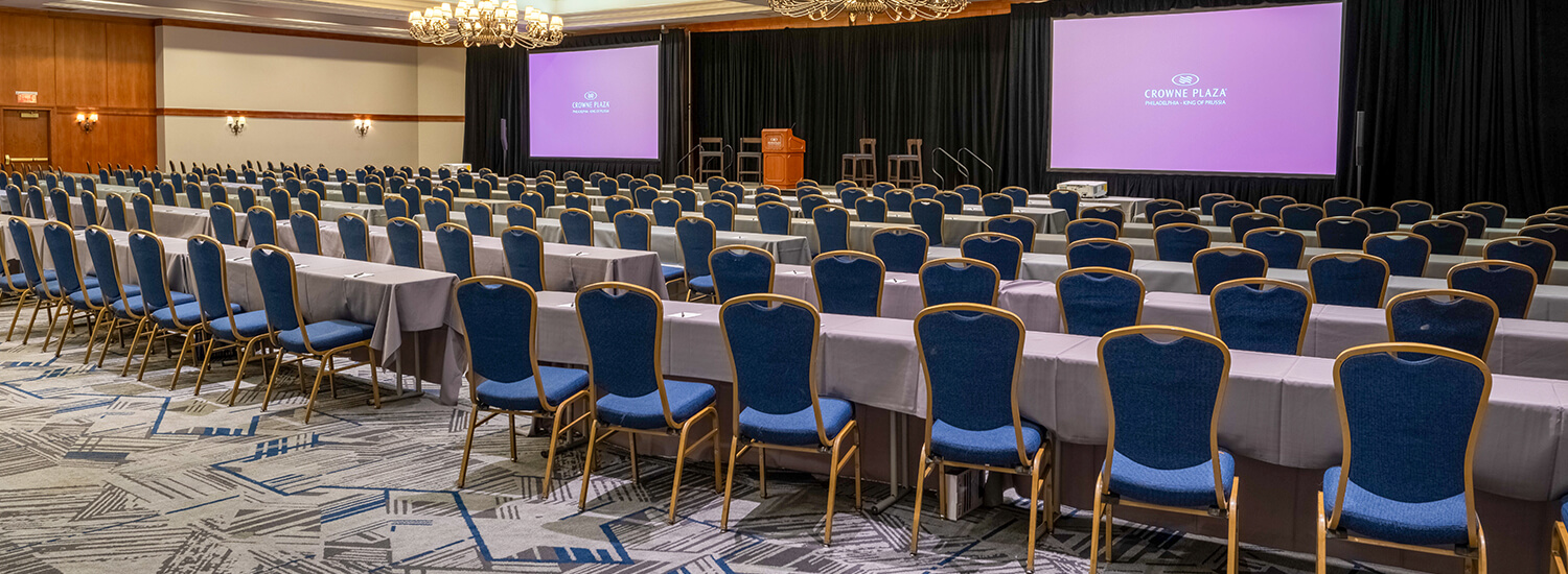 Large ballroom with many rows of tables and chairs with projector screen at from of the room