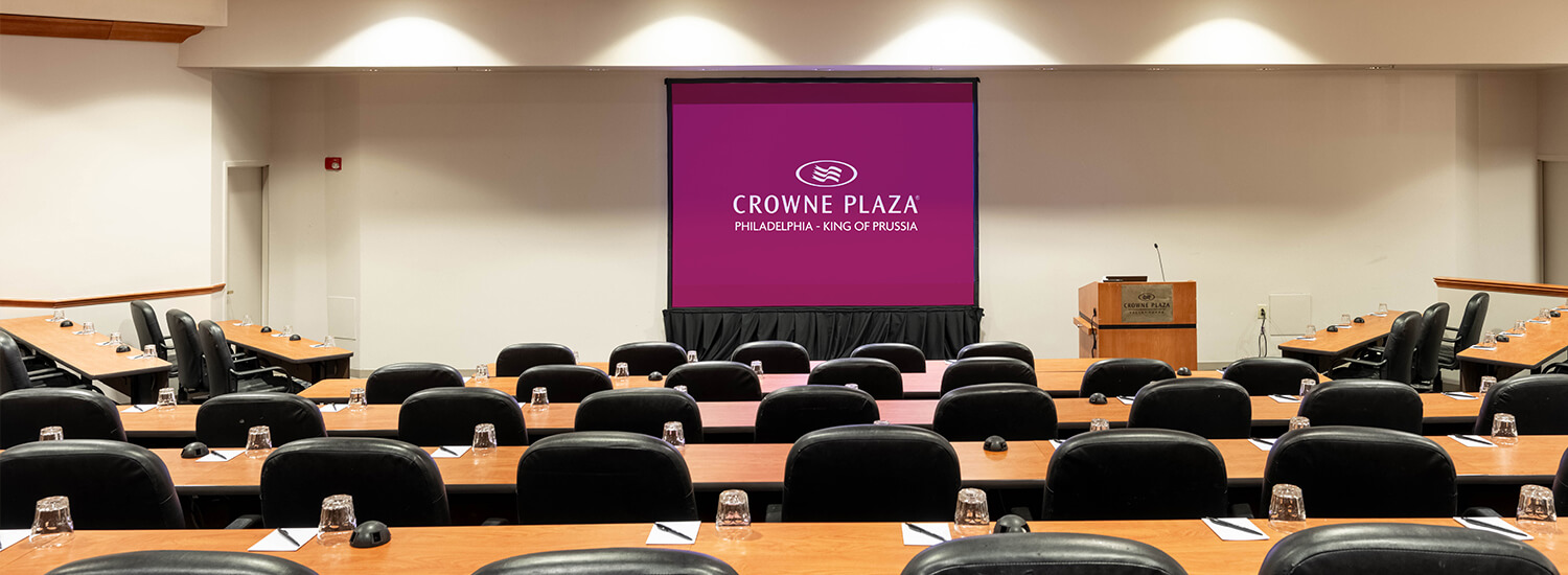 Long brown tables with black chairs and a large projector screen at the front of the room