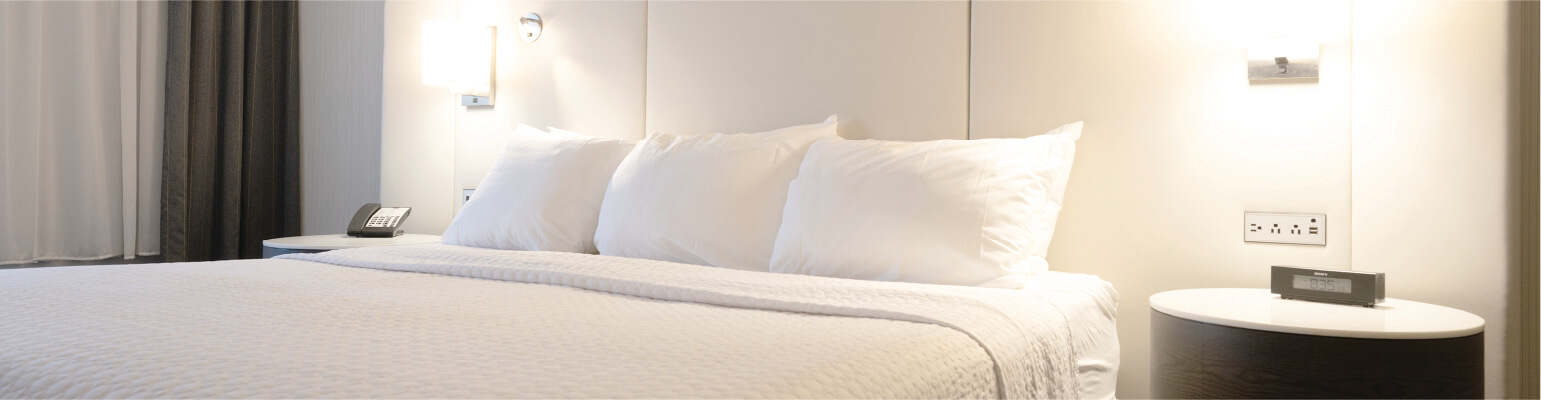 Close up of a bed with 3 pillows and white linens with end tables next to them, one with a phone and the other with an alarm clock on it